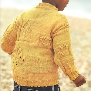 Puede incluir: Un suéter de punto amarillo mostaza con intrincados patrones florales y geométricos. El suéter presenta detalles texturizados, incluyendo bolitas en relieve y diseños en forma de hojas. Las mangas y el cuerpo están adornados con varios patrones de puntadas, creando una prenda visualmente atractiva.