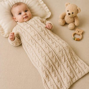 May include: A baby wearing a cream-colored, long-sleeved cable knit sleeping bag with a front zipper closure. The baby rests on a matching cream pillow next to a light brown plush teddy bear and a small wooden bear-shaped rattle toy.