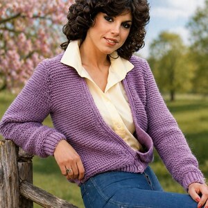 May include: A lavender knitted cardigan with long sleeves and a V-neck, worn over a light yellow collared shirt and blue jeans. The cardigan has a ribbed texture and a single button closure. The background features a wooden fence and a blurred outdoor scene.