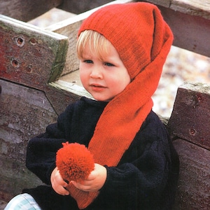 May include: A bright orange knit hat with a long, pointed tail and a matching orange pom-pom attached to the end. The hat features a ribbed texture and is worn over a dark navy blue long-sleeved sweater with a visible knit pattern.