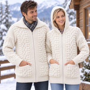 May include: Two cream-colored cable knit sweaters with full-length front zippers and two front pockets. One sweater features a wide collar, while the other includes an attached hood. Both garments are crafted with intricate textured patterns.
