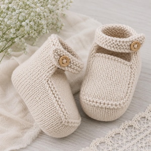 May include: A pair of cream-colored knit baby booties featuring a textured garter stitch pattern, a rounded toe, and a side strap secured with a single light brown wooden button on each shoe. The booties are crafted from a soft, thick yarn material.