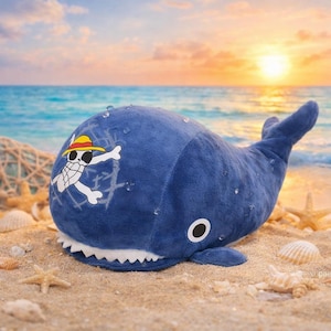 May include: A plush, blue whale toy with a white and yellow pirate logo on its side, resting on a sandy beach with seashells and starfish. The whale has a white, toothy grin and a black and white eye.
