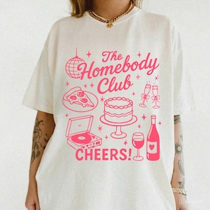 May include: Off-white t-shirt featuring a pink graphic. The design includes the text "The Homebody Club" with illustrations of a disco ball, pizza, cake, champagne glasses, a record player, a wine bottle, and the word "CHEERS!"