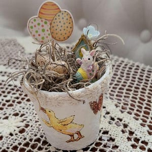 May include: A small, white, decorative pot with an Easter theme. The pot features a yellow duckling and a butterfly design. Inside, there's a nest with speckled eggs, a bunny, and a decorative egg-shaped sign. The pot is filled with dried twigs.