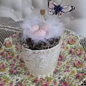 May include: A decorative Easter-themed pot with a nest of white feathers, speckled pink eggs, and a butterfly topper. The pot is cream-colored with lace detailing and sits on a floral patterned tray, creating a charming spring display.