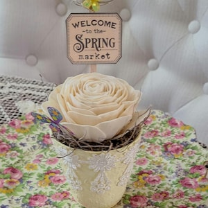 May include: A cream-colored rose in a yellow pot with a lace design, set on a floral tray. A sign that says "Welcome to the Spring Market" is attached to the top of the rose. A decorative butterfly is on the pot.