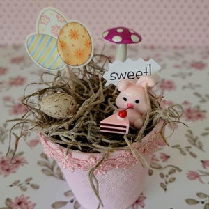May include: A pink decorative pot with a lace trim, filled with dried moss and Easter-themed decorations. Includes a pink bunny, a slice of cake, a mushroom, and egg-shaped cutouts. The word "sweet!" is on a small sign.