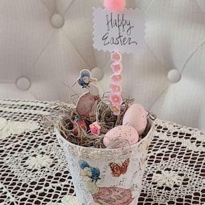 May include: A decorative Easter basket with a duck illustration, pink eggs, and a sign that says "Happy Easter." The basket is filled with decorative grass and small pink flowers. A pink pom-pom tops the sign.