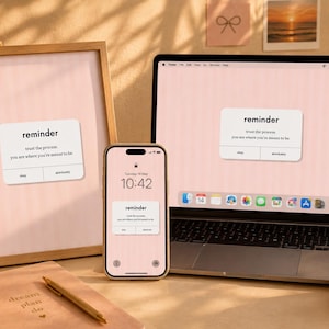 Può includere: Uno sfondo digitale rosa chiaro visualizzato sullo schermo di un portatile, uno smartphone e una stampa incorniciata. Ogni dispositivo mostra una finestra pop-up bianca con il testo reminder, trust the process, you are where you're meant to be, e due pulsanti etichettati okay e absolutely.