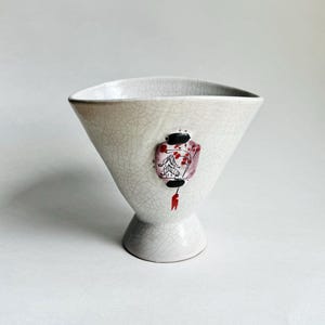 May include: A white, triangular ceramic vase with a pedestal base. The vase features a hand-painted, decorative lantern design in shades of red, black, and pink. The surface has a crackle glaze.