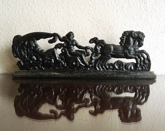May include: A black metal sculpture depicting a chariot scene with a woman, horses, and waves. The sculpture is detailed and has a reflective surface below it. The overall theme is classical art.