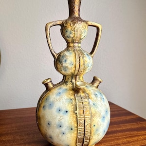 May include: A vintage ceramic vase with a unique shape and design. The vase features a bulbous base with three spouts, a midsection with handles, and a slender neck. The color scheme includes shades of gold, blue, and cream, with a textured surface and decorative detailing.