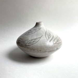 May include: A small, off-white ceramic vase with a rounded body and a narrow neck. The vase features a leaf pattern in a muted gray color. The handmade pottery has a textured surface and is suitable for home decor.