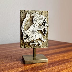 May include: A decorative Sagittarius zodiac sign sculpture. The off-white figure of an archer with a bow and arrow is set against a textured, rectangular background. The word "SAGITTARIVS" is written on a banner. The sculpture is mounted on a brass stand.