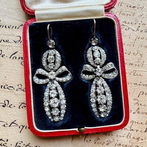 May include: A pair of silver-toned drop earrings featuring a bow-shaped design encrusted with clear, round-cut stones. The earrings are displayed inside a rectangular red jewelry box with a dark blue velvet interior lining and a cream-colored lid.