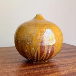May include: A spherical ceramic vase with a narrow opening at the top. The surface features a glossy mustard yellow glaze with vertical drip patterns revealing a deep reddish-brown base color underneath, creating a layered and textured appearance.