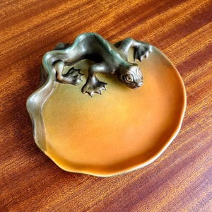 May include: A round ceramic dish featuring a sculpted lizard figure attached to the rim. The dish has a smooth, gradient finish transitioning from a deep forest green at the edges to a warm, vibrant orange in the center, with a glossy surface.