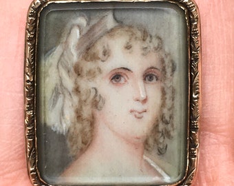 May include: A rectangular gold-toned brooch featuring a miniature portrait of a woman. The portrait depicts a woman with light skin, curly blonde hair, and a white head covering. The brooch has a decorative, engraved border.