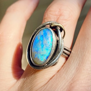 May include: Oval opal ring with a silver band and a gold accent. The opal displays a vibrant play of color, with blues and hints of other colors. The ring has a unique, artistic design.