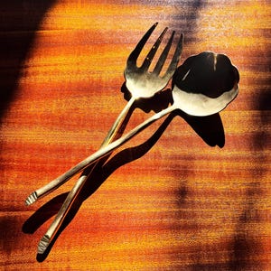 May include: A gold-colored serving fork and spoon set. The fork has four tines, and the spoon has a scalloped bowl. The set is lying on a wooden surface with a warm, reddish-brown tone. The lighting creates long shadows.
