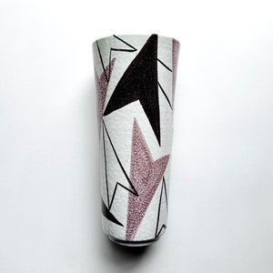 May include: A tall, cylindrical vase with a white background and a geometric pattern. The design features black and mauve-colored angular shapes and lines. The vase has a slightly textured surface.