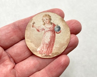 May include: A round, antique illustration of a figure in a pink dress holding a cross and a blue globe. The artwork is hand-painted with delicate details and soft colors, set against a cream-colored background. The image is held in a hand.