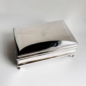 May include: A rectangular silver box with a reflective surface. The lid features an engraved monogram and is bordered by a beaded design. The box rests on small, rounded feet. The bottom edge has engraved text.