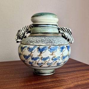 May include: A ceramic jar with a lid, featuring a rounded body decorated with a repeating pattern of blue birds on a cream background. The upper section has a textured cross-hatch design, and the two side handles are sculpted with black and white stripes.