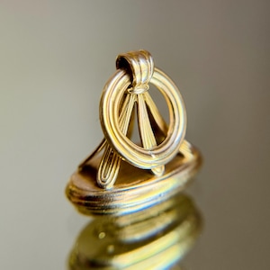 May include: A gold-toned metal wax seal stamp featuring an oval base and a vertical handle design. The handle consists of a circular ring supported by three radiating, fluted struts that connect to the top of the oval base.