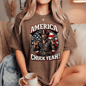 May include: Brown t-shirt featuring a graphic of a man in a cowboy hat holding two guns, with the text "AMERICA" above and "CHUCK YEAH!" below. The design incorporates the American flag.