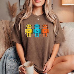 May include: Brown t-shirt featuring three colorful robot graphics. The robots are yellow, blue, and orange, each with a unique binary number: 01, 10, and 11. The shirt is a comfortable, oversized fit.