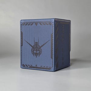 Może przedstawiać: Niebieskie pudełko z teksturowaną powierzchnią i czarną grafiką robota. Pudełko ma prostokątny kształt z zamknięciem na klapę. Posiada czarny tekst z boku i czarną obwódkę.