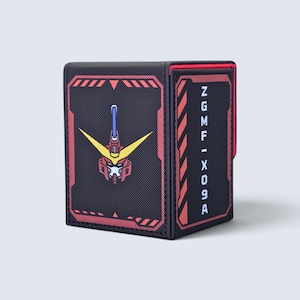 May include: A black deck box with a textured surface featuring a red and yellow robot head graphic on the front panel. The side panel displays red diagonal warning stripes and the vertical white text ZGMF-X09A against a black background.