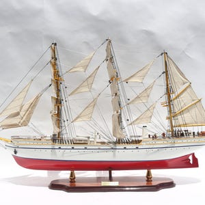 Puede incluir: Maqueta detallada de un velero de tres mástiles con numerosas velas. El barco tiene un casco blanco con una línea de flotación roja y está montado sobre una base de madera. Las velas son beige y el aparejo es visible.