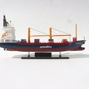 Pode incluir: Modelo de navio de carga, pintado em azul, vermelho e branco, com o nome "Corinna" na proa. O navio apresenta guindastes, contêineres e a palavra "speedline" na lateral. É montado em um suporte preto.