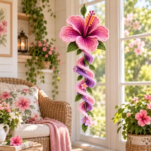 May include: A hanging crochet decoration featuring a large, vibrant pink hibiscus flower with a yellow stamen at the top. Below the flower, a long, spiraling vine in shades of purple and pink is adorned with small green leaves and tiny purple flowers.