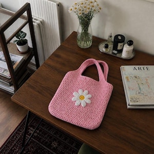 May include: A pink crocheted tote bag with two handles and a large white daisy applique with a yellow center. The bag is sitting on a wooden table. A vase of daisies and a magazine are also on the table.