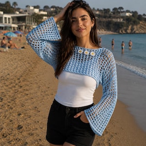 May include: A light blue crochet long-sleeve bolero top featuring a wide mesh pattern and three small white daisy flower appliqués at the neckline. The garment is worn over a plain white tank top paired with black denim shorts.