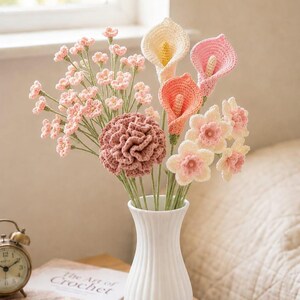 May include: A bouquet of handmade crochet flowers in a white vase. The arrangement includes pink, peach, and cream-colored calla lilies, carnations, and other delicate blossoms. The vase sits on a table with a book and a small clock.