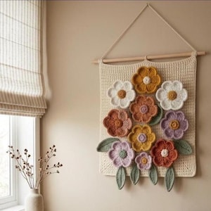 Könnte beinhalten: Ein handgefertigter Wandbehang aus Häkelarbeit mit einem Bouquet aus bunten Blumen. Die Blumen sind in Weiß-, Orange-, Gelb- und Lilatönen gehalten, mit grünen Blättern. Der Wandbehang ist mit einer cremefarbenen Schnur an einer Holzstange befestigt.