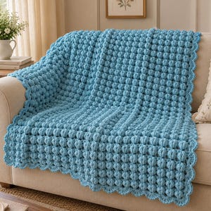 May include: A light blue crochet blanket featuring a repeating bobble stitch pattern throughout the main body. The edges of the rectangular blanket are finished with a decorative scalloped border, creating a textured and uniform appearance.