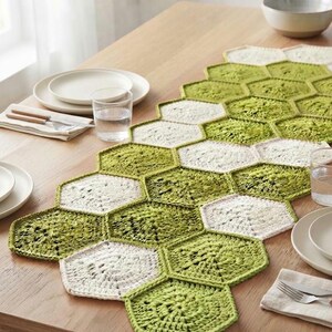 May include: A table runner constructed from interconnected hexagonal crochet motifs in alternating shades of light green and off-white. The textile features a geometric honeycomb pattern, creating a textured surface that spans the length of the table.