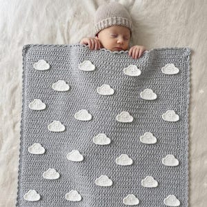 May include: A square, gray crocheted blanket featuring a repeating pattern of small, white, three-dimensional clouds attached to the surface. The blanket has a scalloped edge and is displayed with a beige knit hat resting at the top edge.