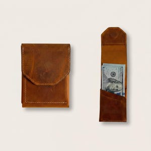 May include: A tan leather wallet with a fold-over flap closure shown in a closed position and an open position containing a one hundred dollar bill. The wallet features visible stitching along the edges and a smooth, matte leather texture.