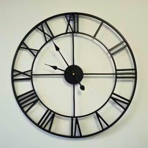 May include: A large, round black metal wall clock with Roman numerals. The clock face is white, and the hands are black. The clock has a minimalist design, suitable for home decor. The clock's diameter is approximately 61 cm.