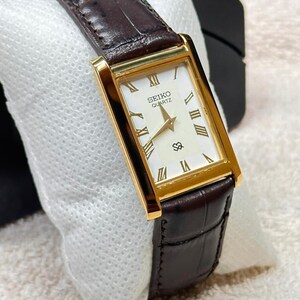 May include: A rectangular Seiko Quartz wristwatch featuring a gold-tone case, a white dial with gold-tone Roman numeral hour markers, and a dark brown leather strap with a textured crocodile-embossed pattern and gold-tone hands.