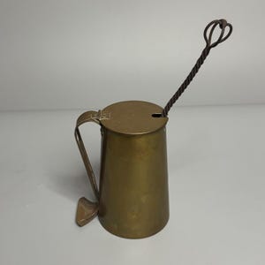 May include: A cylindrical brass vessel with a flat hinged lid and a curved handle. A long, thin, twisted metal rod with a looped top extends through a small notch in the lid. The brass has a muted, aged finish, and the metal rod is dark and textured.