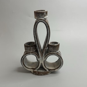 May include: A ceramic candle holder with a unique design. The holder features three cylindrical candle cups and a central loop. The color is a mix of brown and white, with a textured appearance. The base is round.
