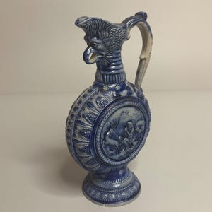 May include: A blue and white ceramic pitcher featuring a sculpted griffin head at the spout and a circular relief panel on the body depicting a figure with a deer in a forest setting. The vessel has a tall handle and a decorative pedestal base.